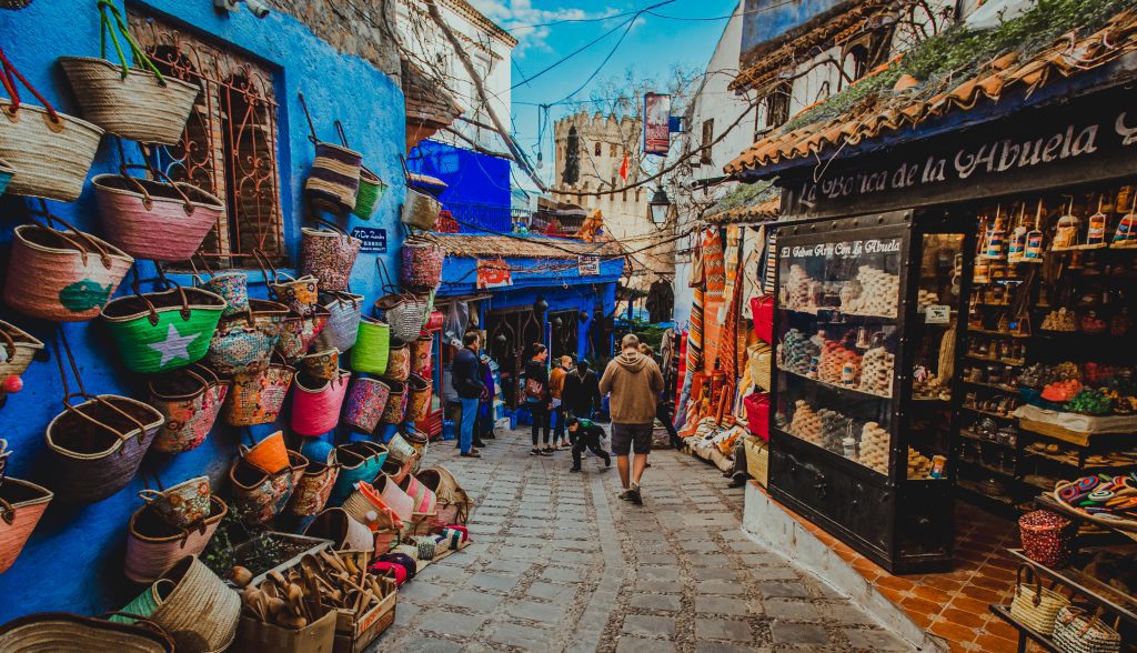 souvenir shops with dresses, carpets and handcrafted stuff and people walking in Chefchaouen’s Ensemble Artisanal on Place el Makhzen